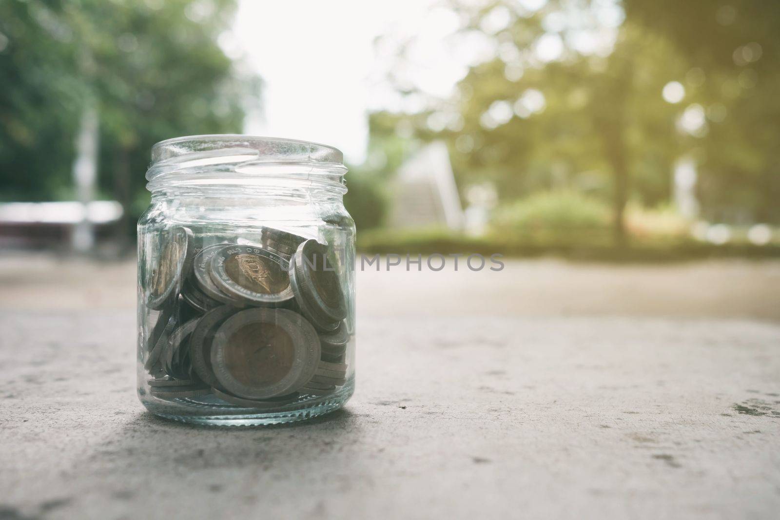 Coins in Glass Jar in Park with Light Leak. Translation Text on Coin is "Thailand, 10 Bath". (Baht is Currency of Thailand)