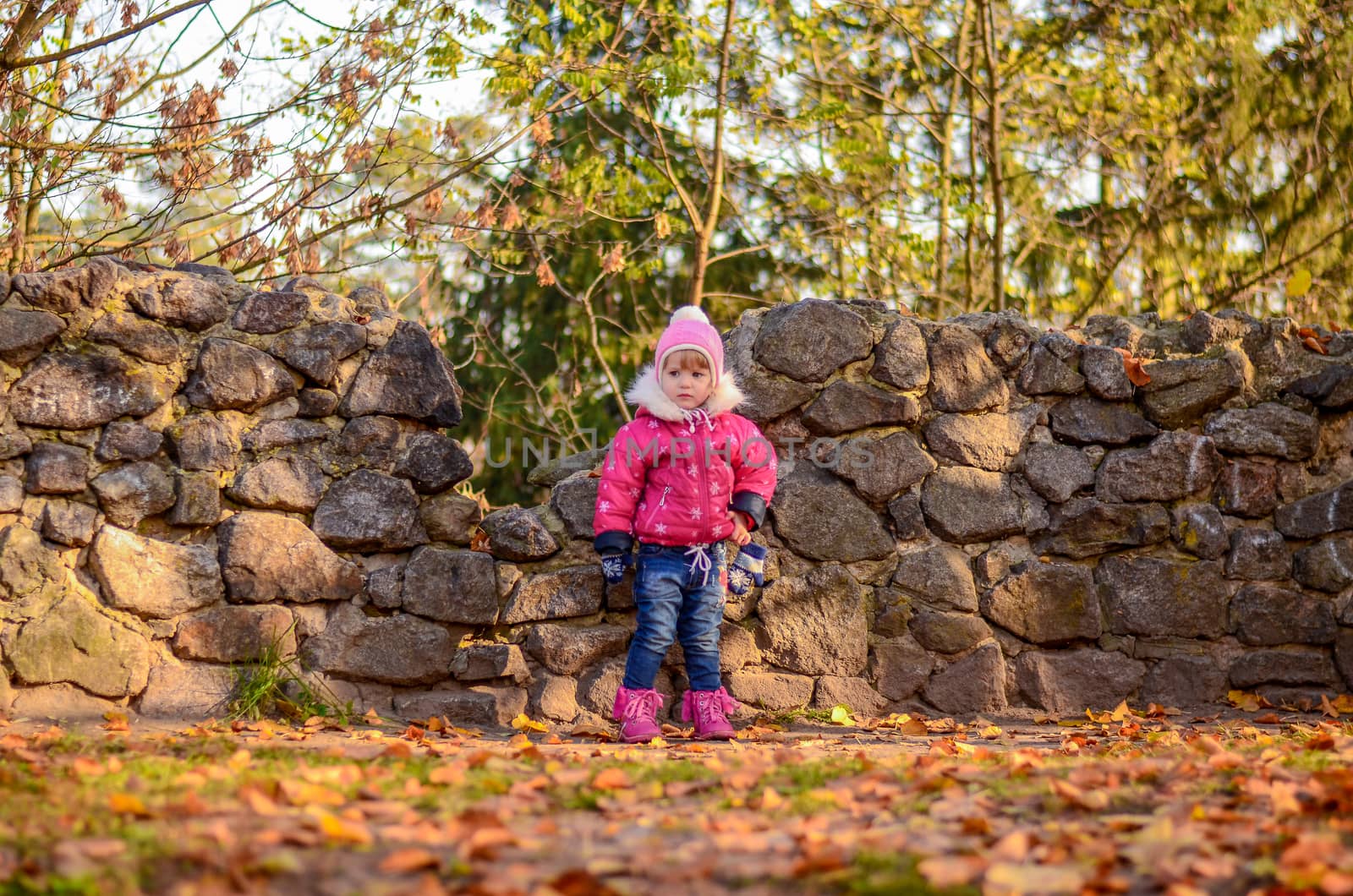Little girl in winter clothes stands on stone wall background by chernobrovin