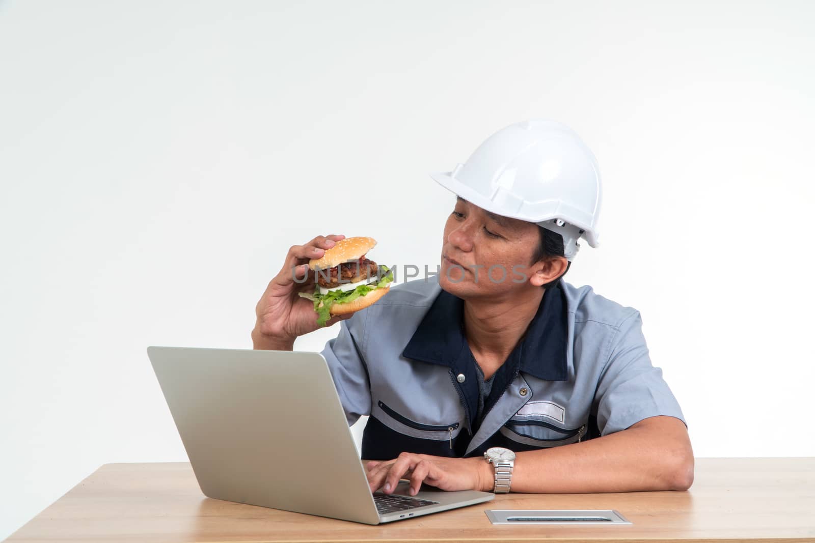 Asian man worker smiling and holding a hamburger in the living room While working from home, hem very happy and enjoy to eat fast food. Concept of binge eating disorder (BED). by PattyPhoto