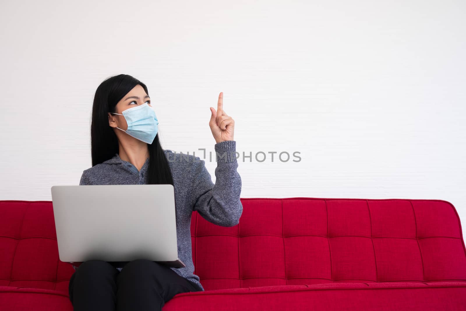 Asian woman wearing a face mask and using a laptop on the sofa for working from home and Pointed to the top. New lifestyle normal during a quarantine. Concept of stay home. by PattyPhoto