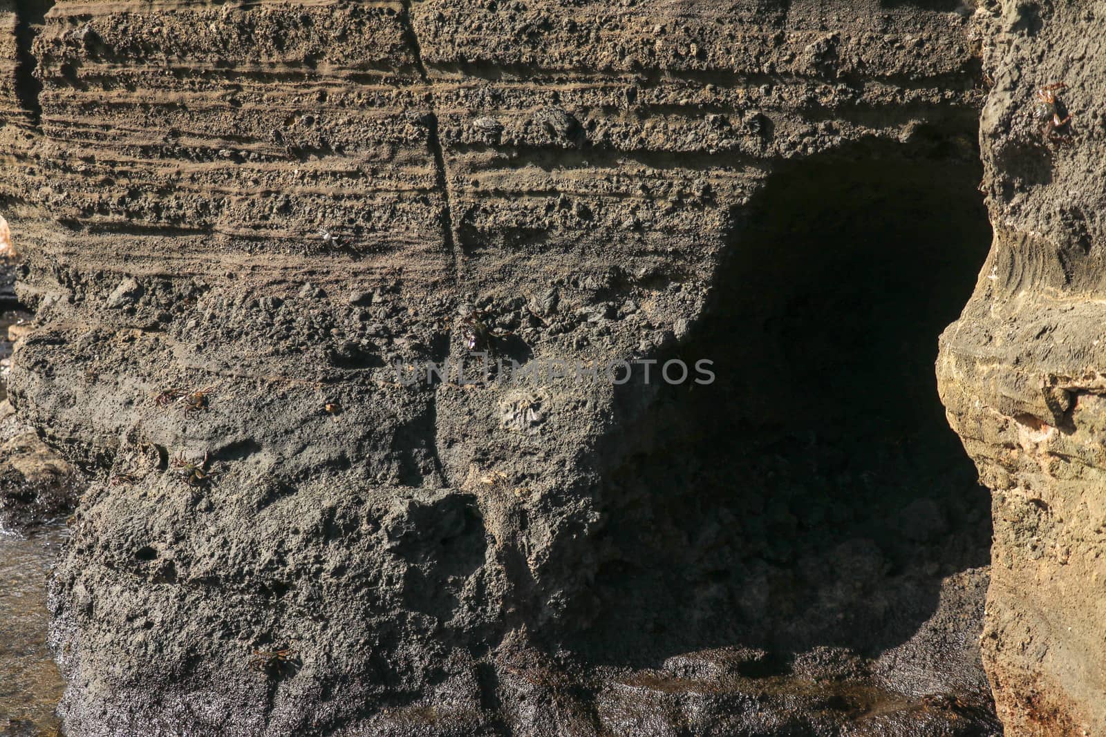 Dark rock of volcanic lava on the Indian Ocean coast of Bali. A small cave excavated into a cliff wall by a sea surf. Sun rays illuminate the rough wall of a rock cliff.