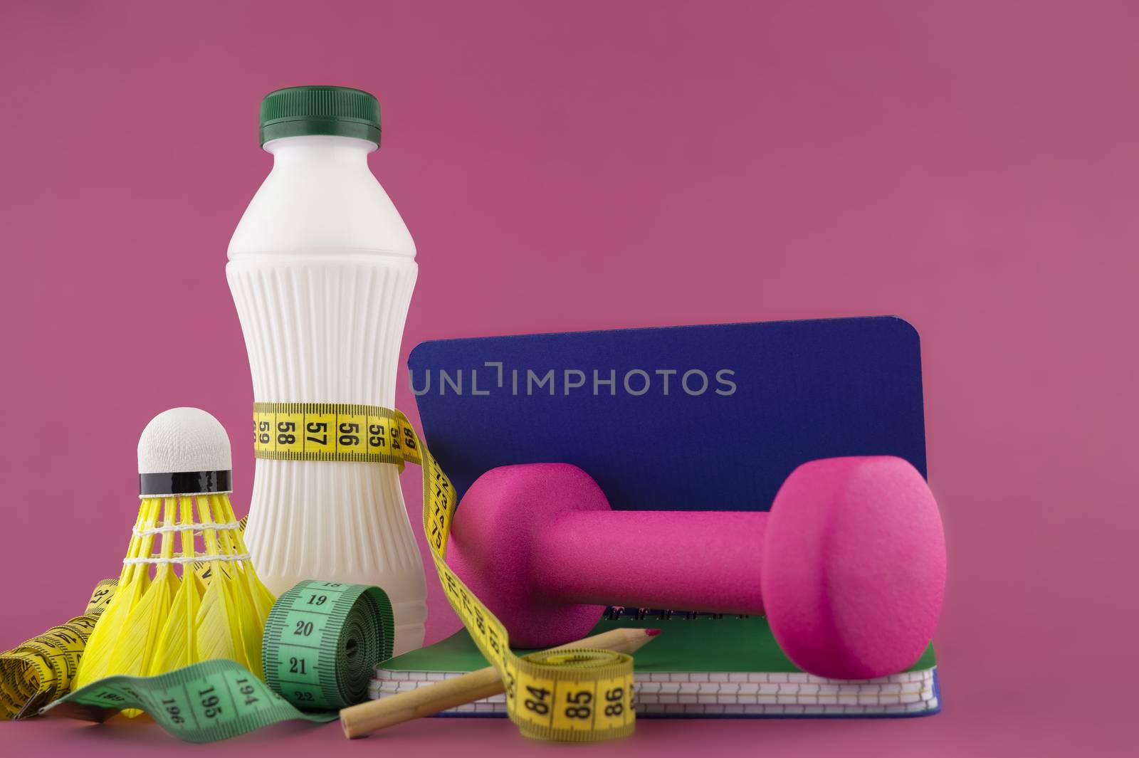 Sport weight loss and fitness concept with a shuttlecock, dumbbell weight and tape measure wound round a white plastic bottle with a slim line shape, open notebook with pen on a bright pink background