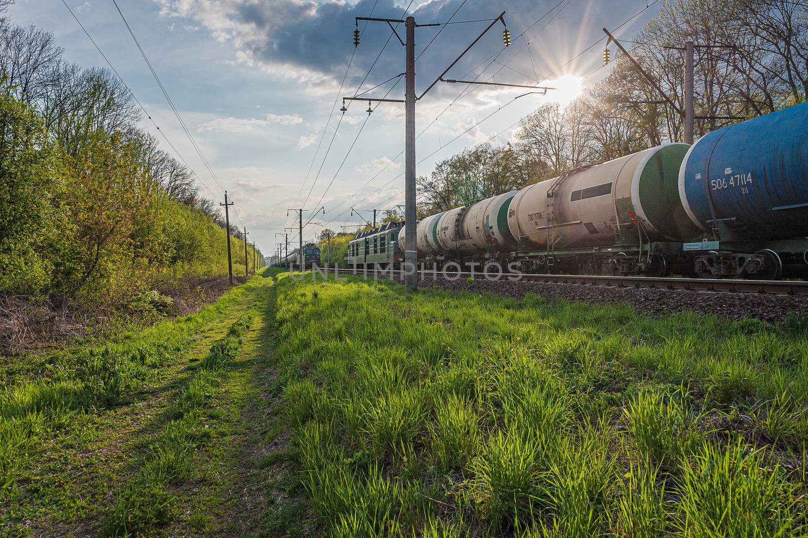 two trains with tank wagons move on railway tracks towards each other at sunset near green grass and trees