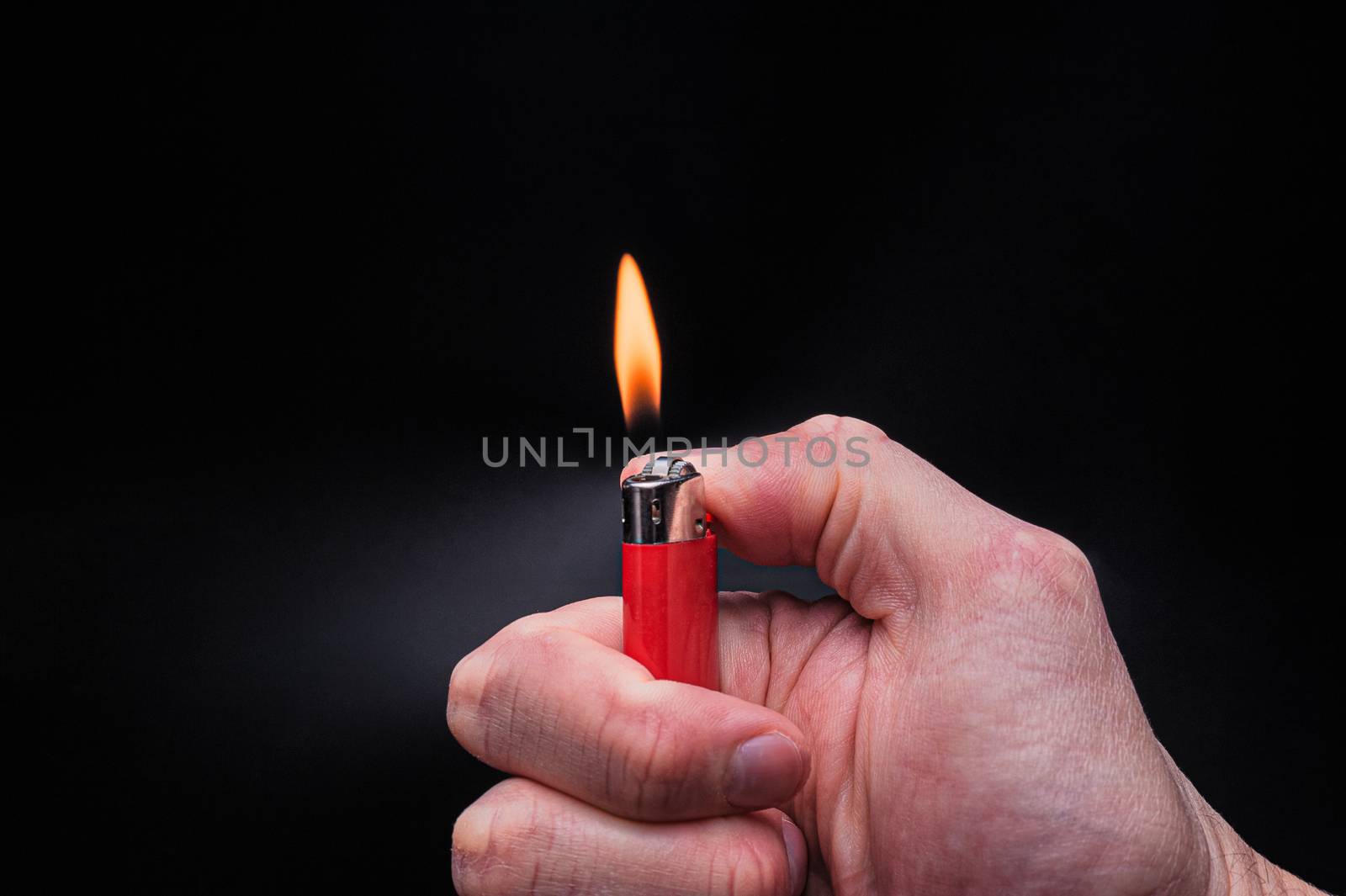 European white mans hand holds a burning red ignited plastic gas lighter with fire on an isolated black background