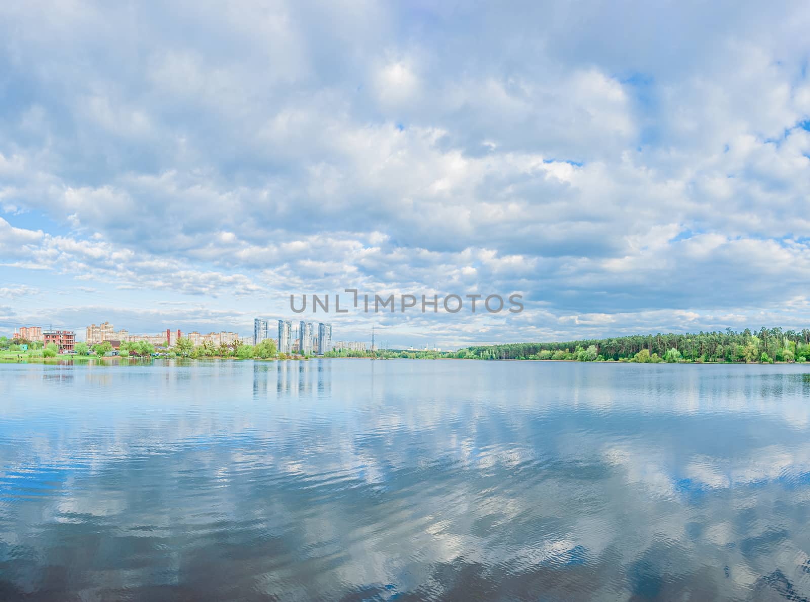 Large blue lake and sky with beautiful clouds with green forest, houses and skyscrapers on the shore. Panorama landscape