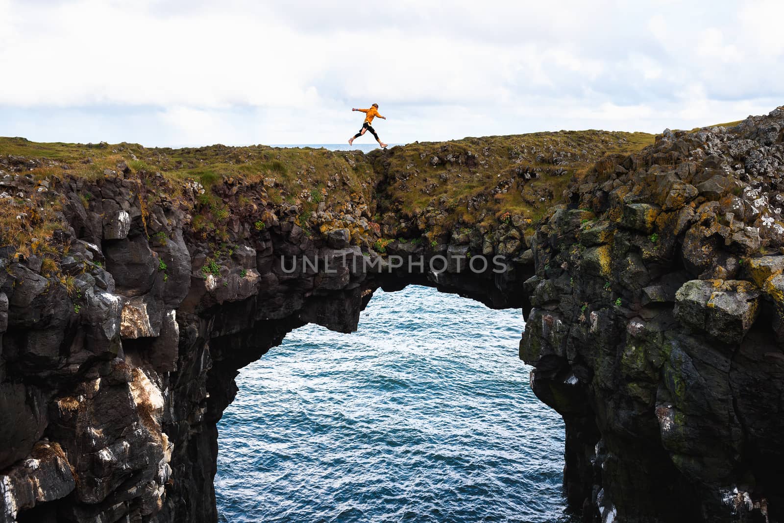 Tourist jumps over a natural rock bridge in Arnarstapi, Iceland by nickfox