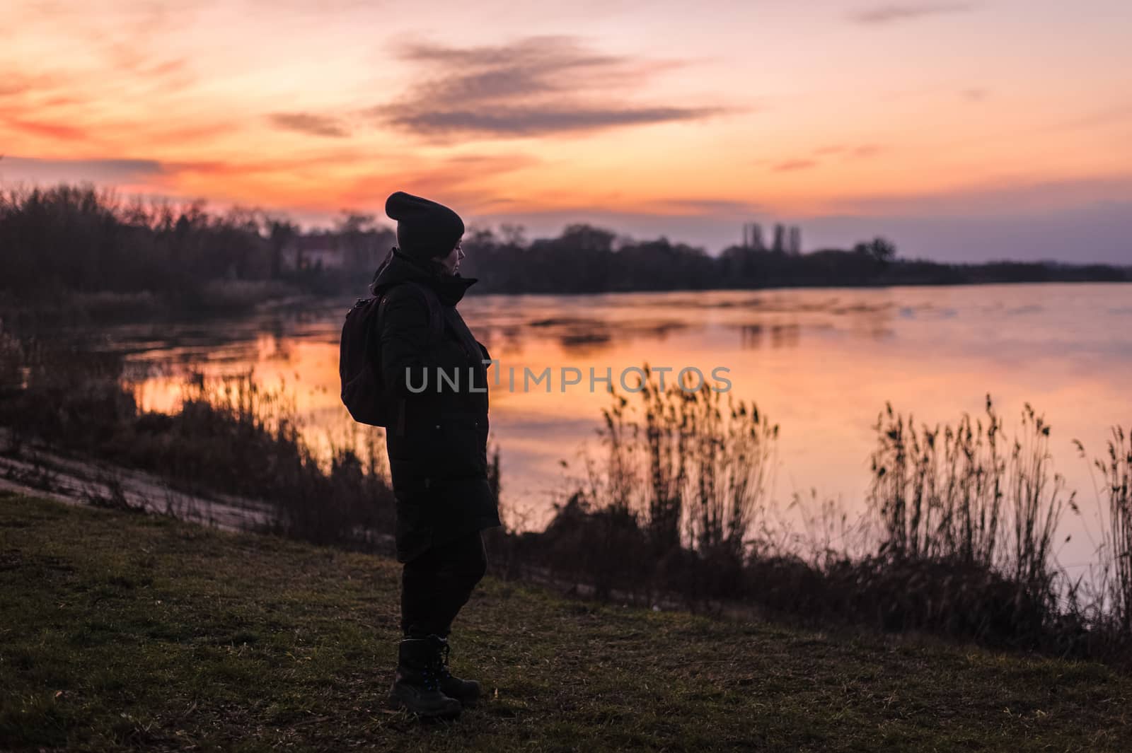 girl in winter clothes stands on a lake at sunset