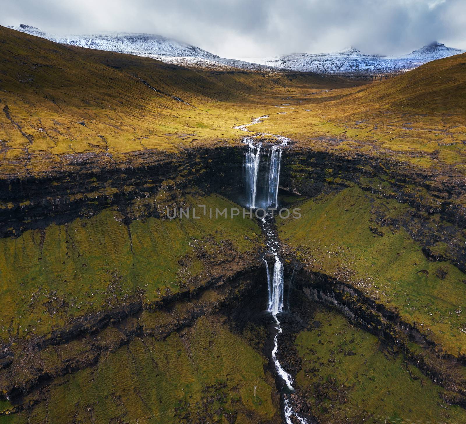 Aerial view of the Fossa Waterfall on Bordoy island with snow covered mountains in the background. This is the highest waterfall in the Faroe Islands, situated in wild scandinavian scenery.