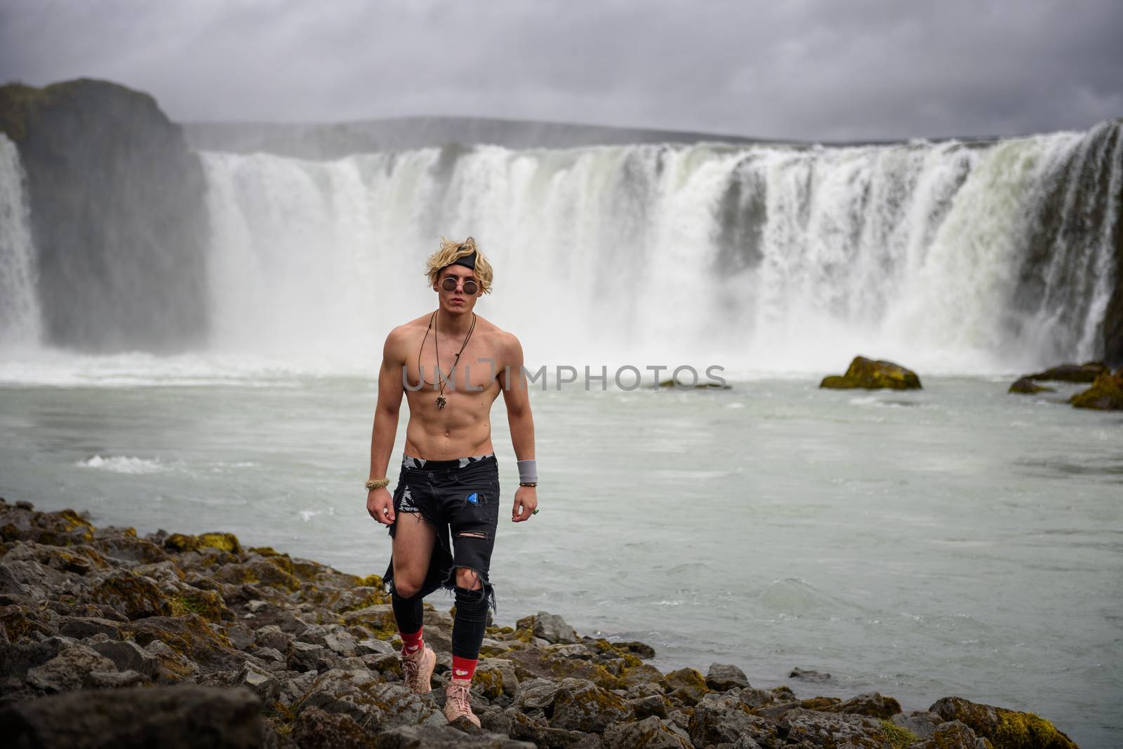 Shirtless and muscular teenage boy walks around the Godafoss waterfall in Iceland. Godafoss means the waterfall of the gods in icelandic.