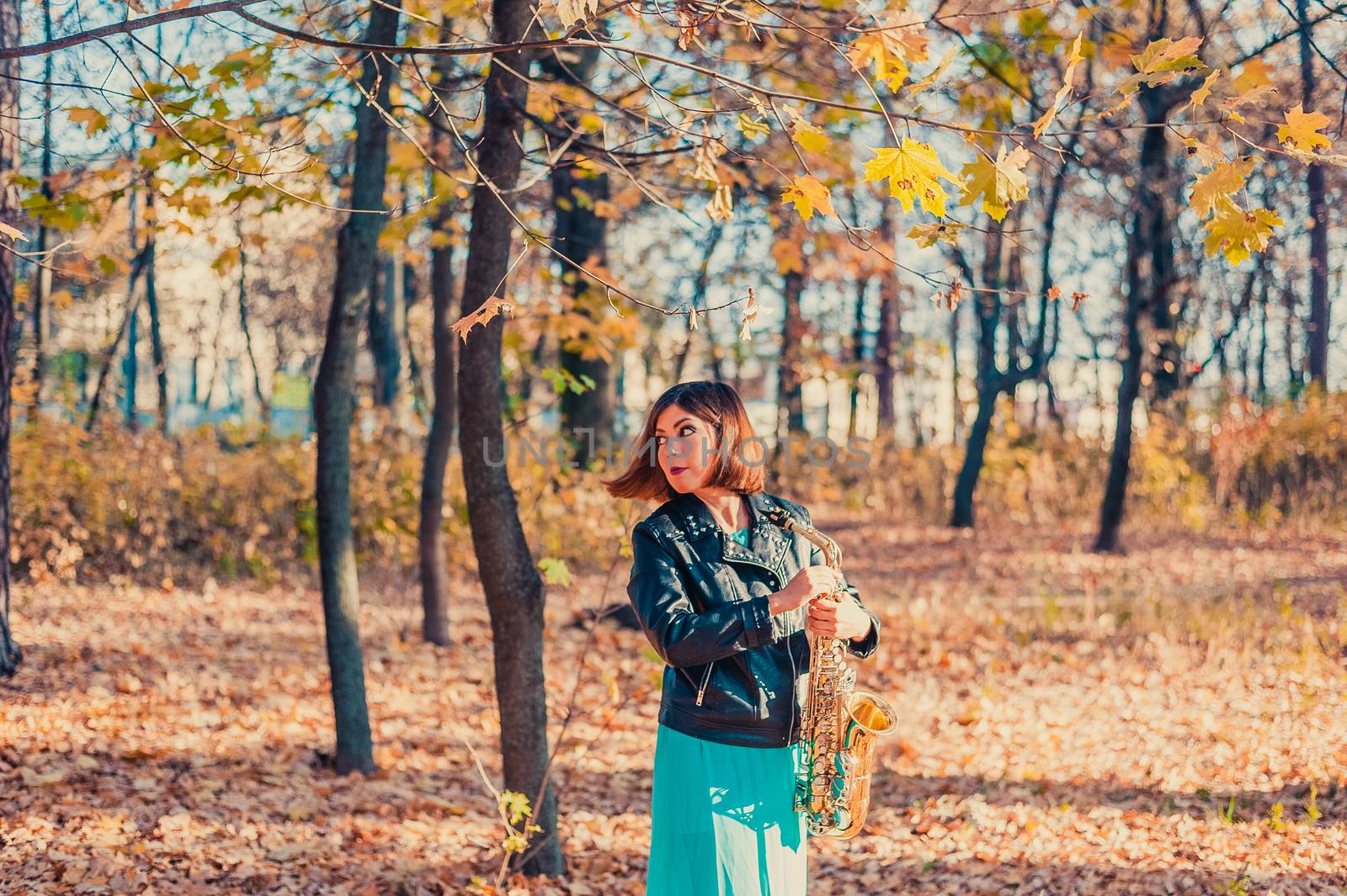 pretty dark-haired girl in a black jacket and blue skirt with a saxophone in a yellow autumn park by chernobrovin