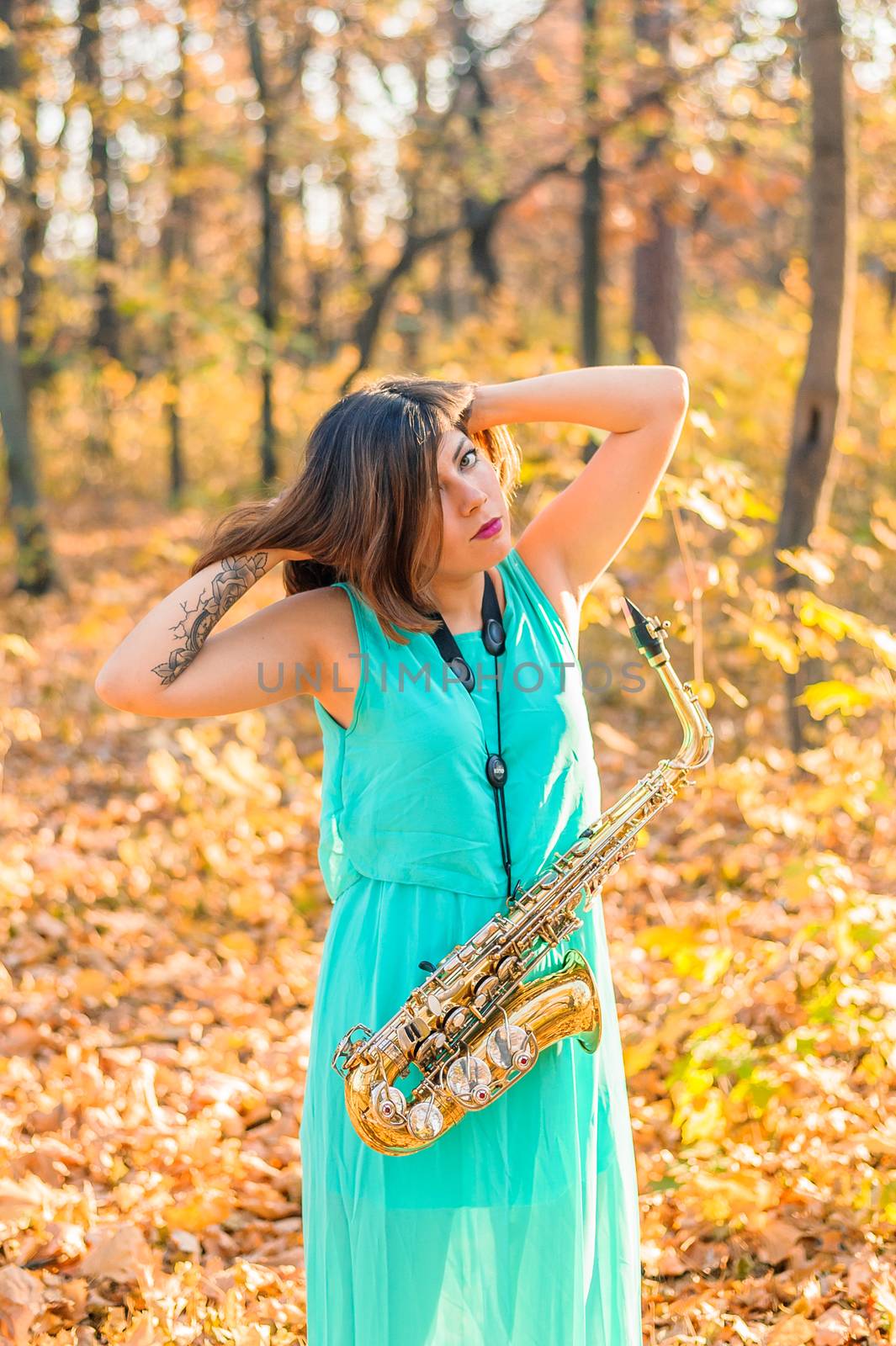 lovely brunette girl in a long blue dress and alto saxophone posing in a yellow autumn park, raising hands to head