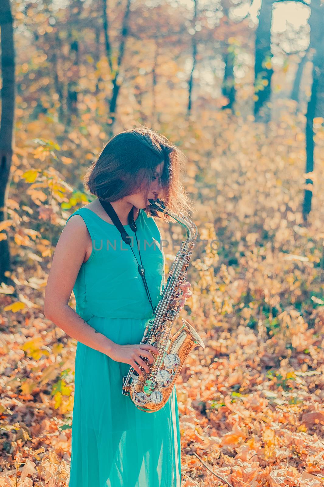 cute young brunette girl in a long blue dress plays the yellow saxophone in the autumn forest