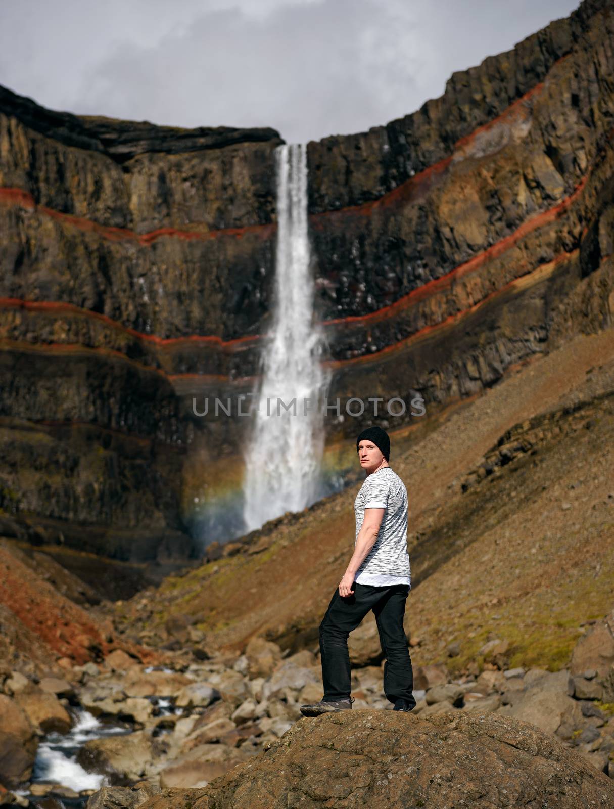Tourist looking at the Hengifoss waterfall in Iceland. Hengifoss is the third highest waterfall in Iceland and is surrounded by basaltic strata with red layers of clay between the basaltic layers.