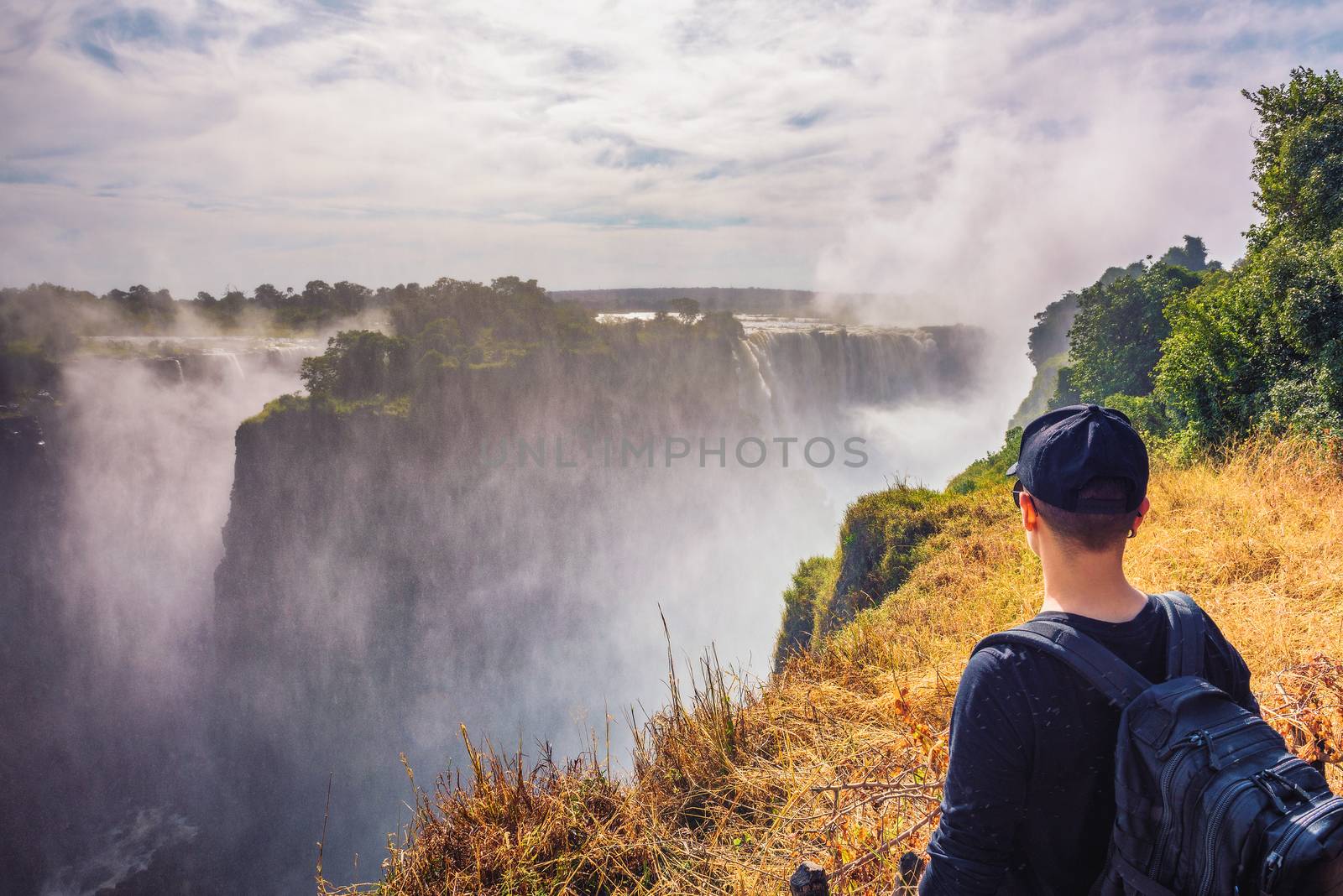 Tourist with a backpack looks at the Victoria Falls on Zambezi River located at the border of Zambia and Zimbabwe, the largest waterfall in the world.