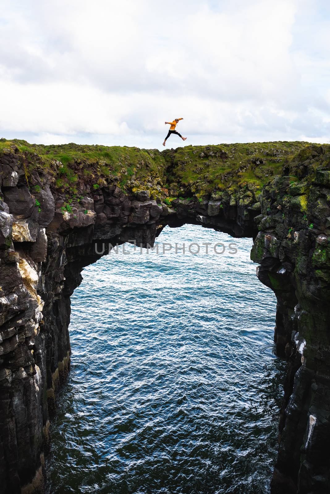 Tourist jumps over a natural rock bridge in Arnarstapi, Iceland by nickfox