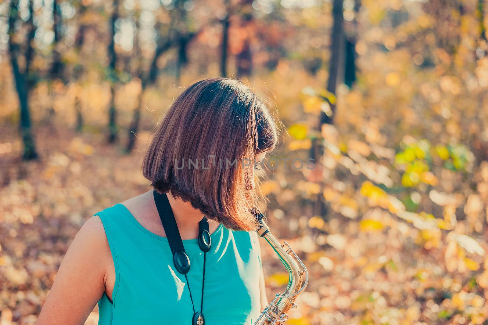 sideways from close up a brunette woman in a blue dress plays the saxophone in a yellow autumn park