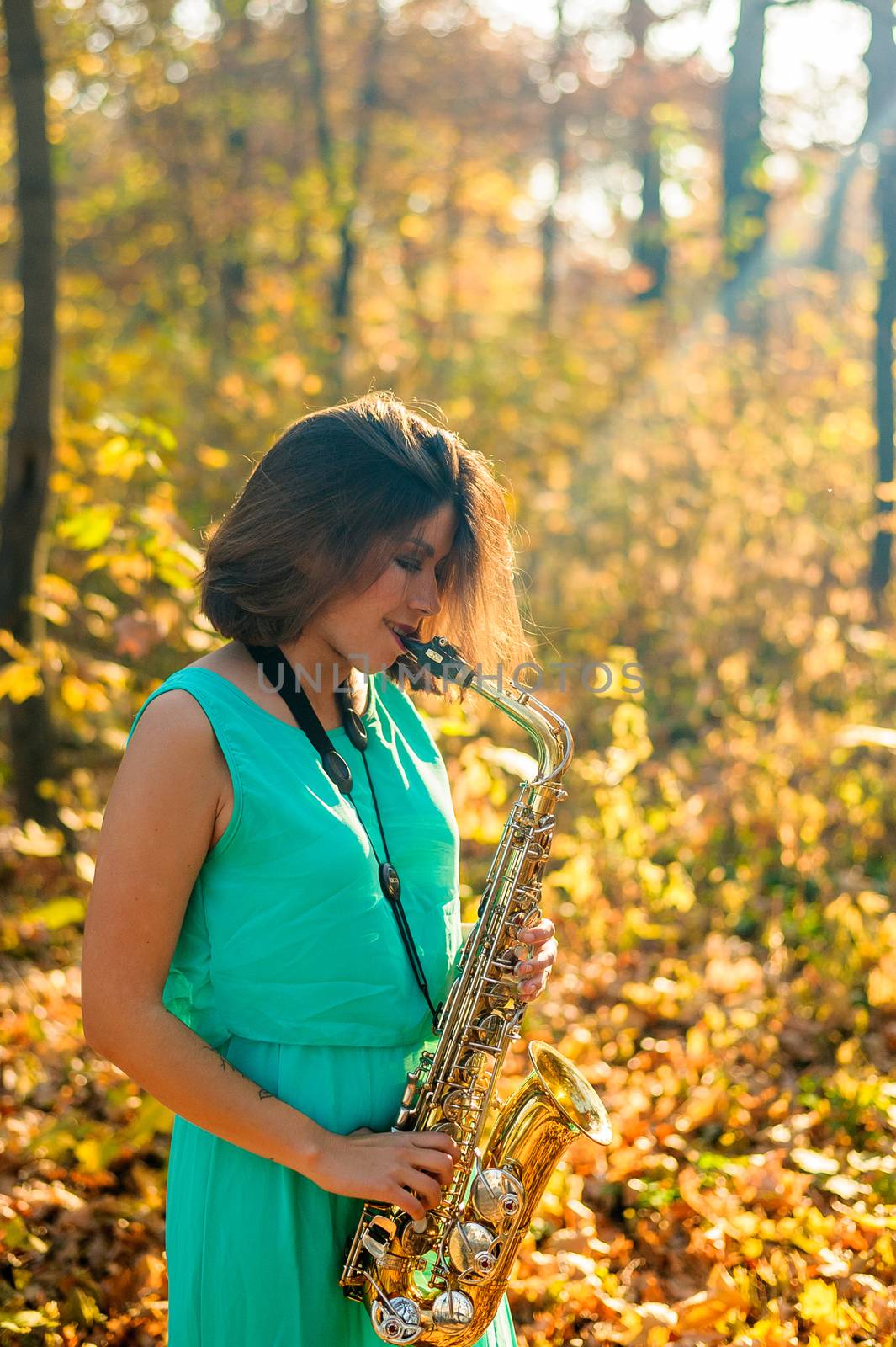 cute black-haired girl in a blue dress plays the yellow saxophone in the autumn forest by chernobrovin