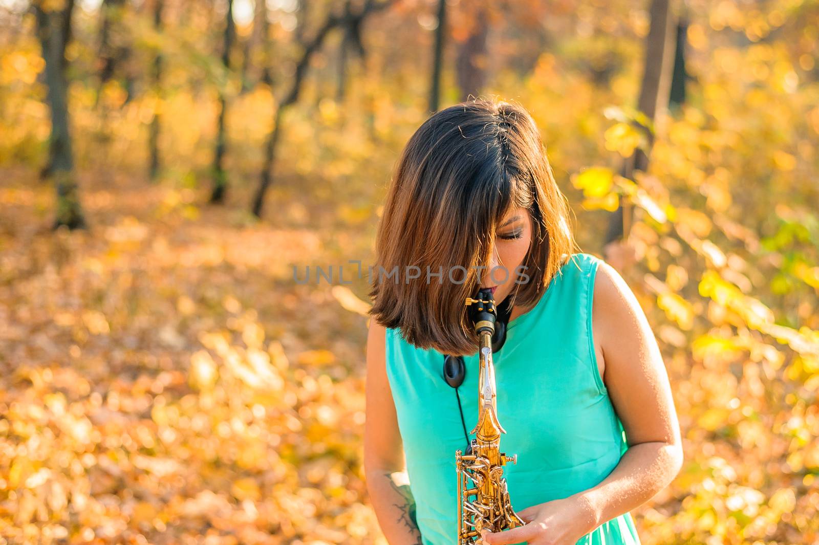 Young brunette girl with hair covering her face plays the saxophone, standing in blue clothes in a yellow autumn park
