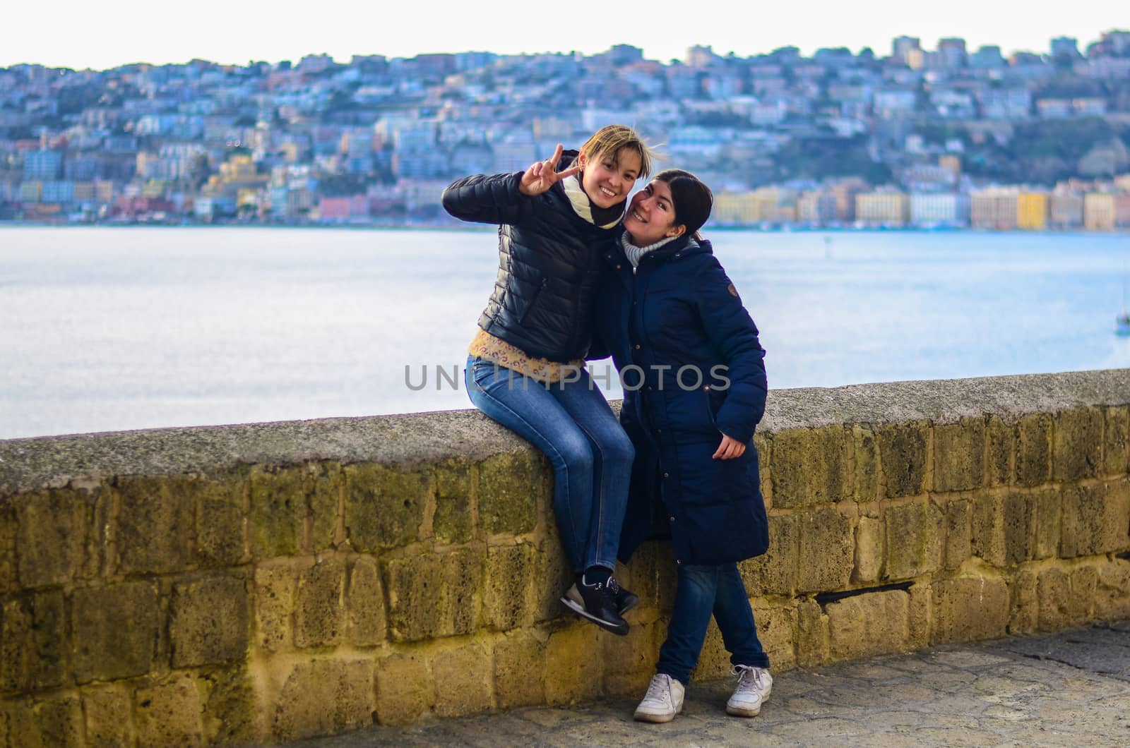 Two young smiling women on the background of the city at sunset. Blue sea ​​in the background by chernobrovin