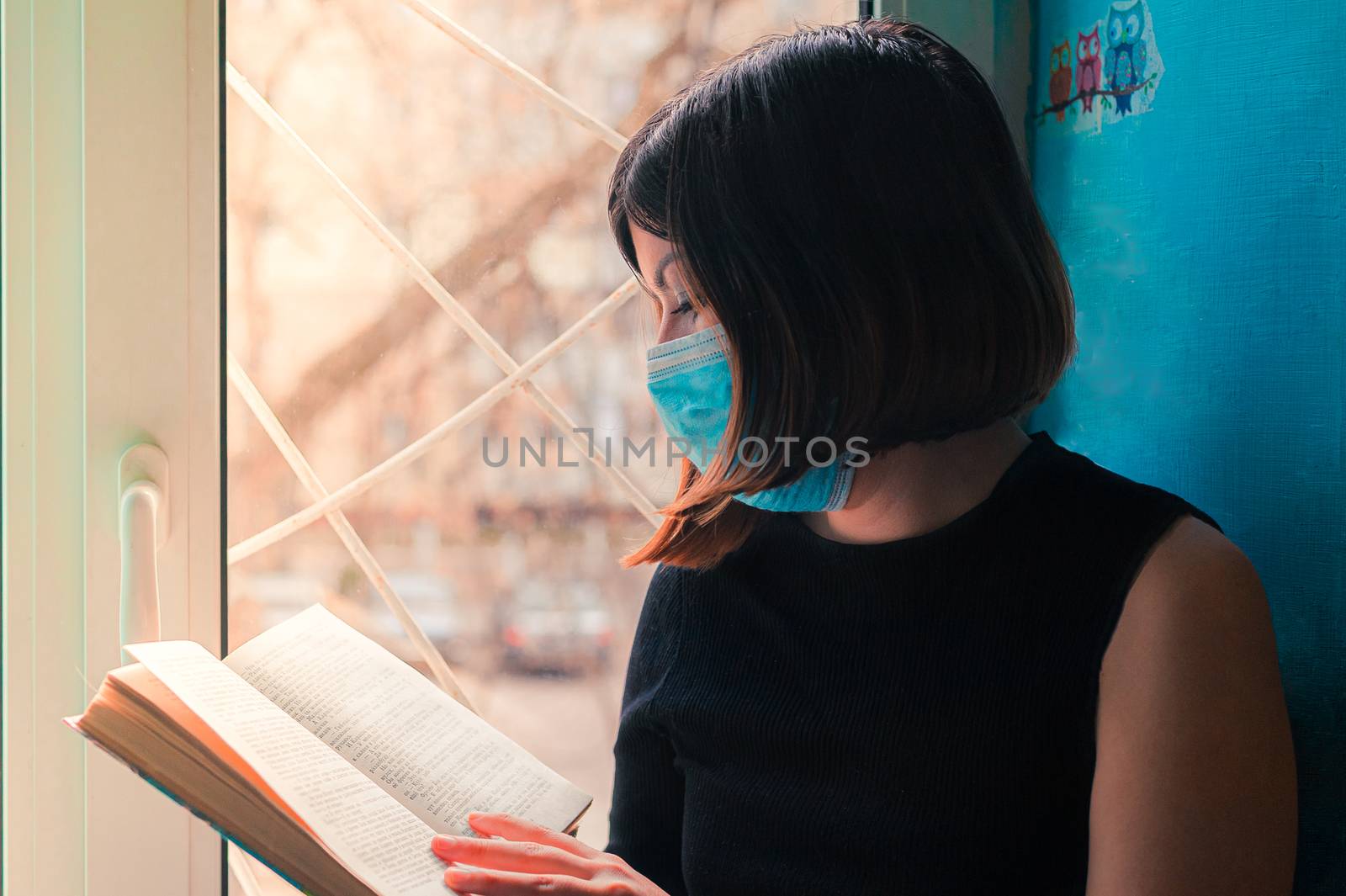 a dark-haired girl in a black dress sits in a surgical mask at the hospital window and reads a book