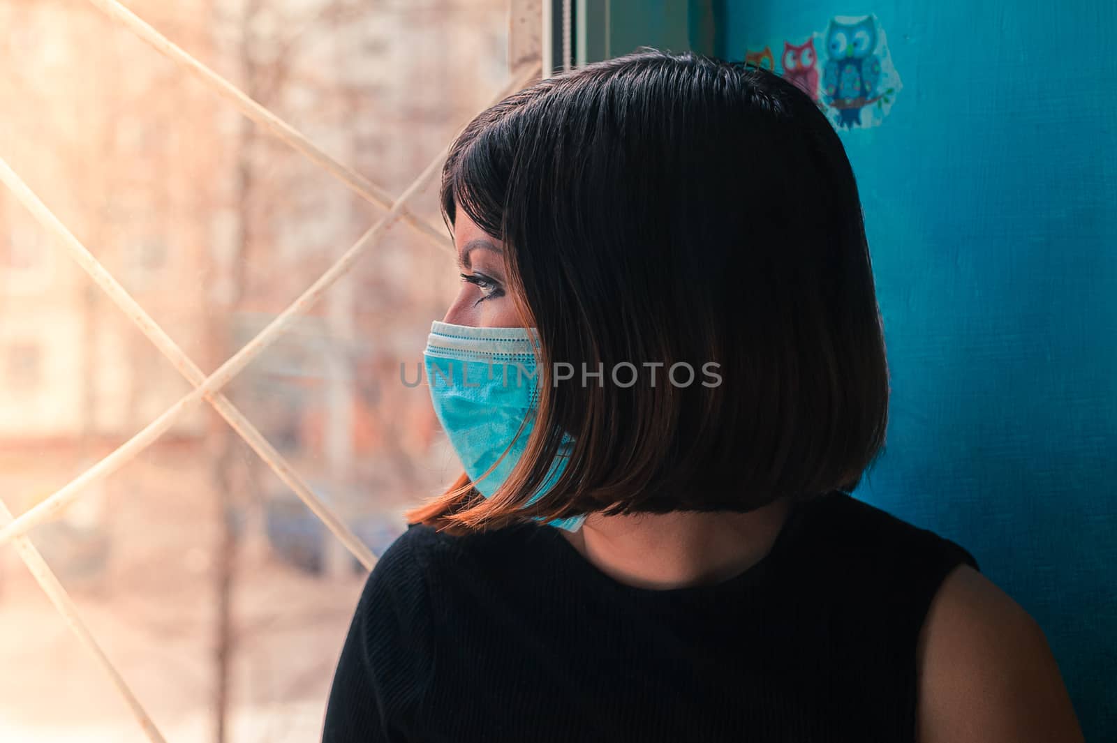 brunette girl in a black dress sits in a surgical  mask sits on a hospital window and looks outside