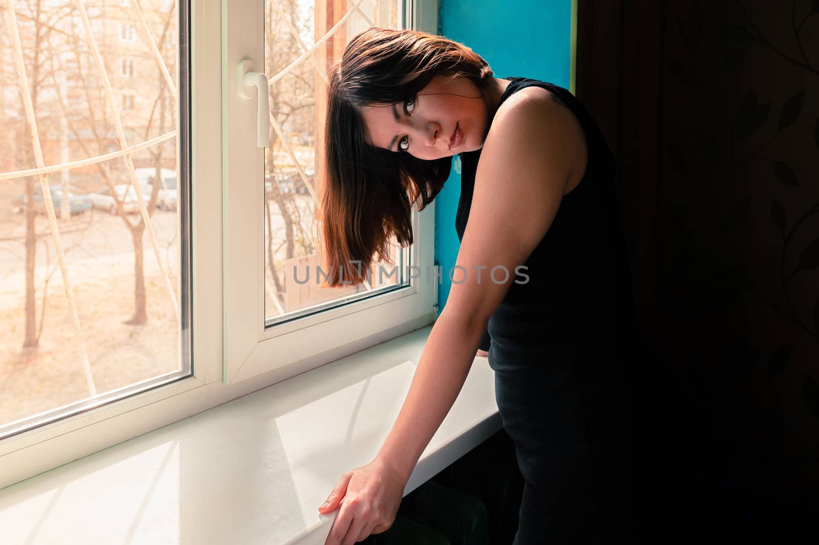 young beautiful brunette girl in a black dress leaned on a windowsill near a barred window by chernobrovin