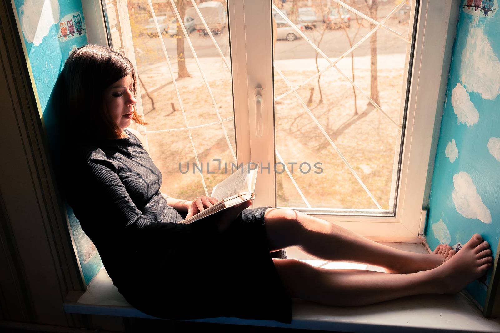 charming young brunette woman in a long black dress sits on a windowsill by the window and reads a book by chernobrovin