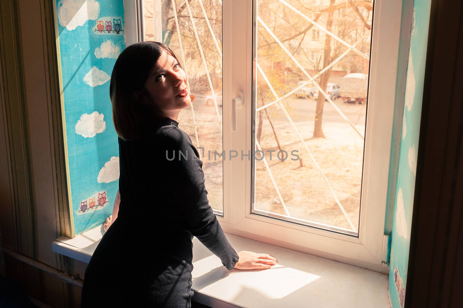 unusual beautiful young girl with black hair in a long tight dark dress stands by the window, leaning on a windowsill