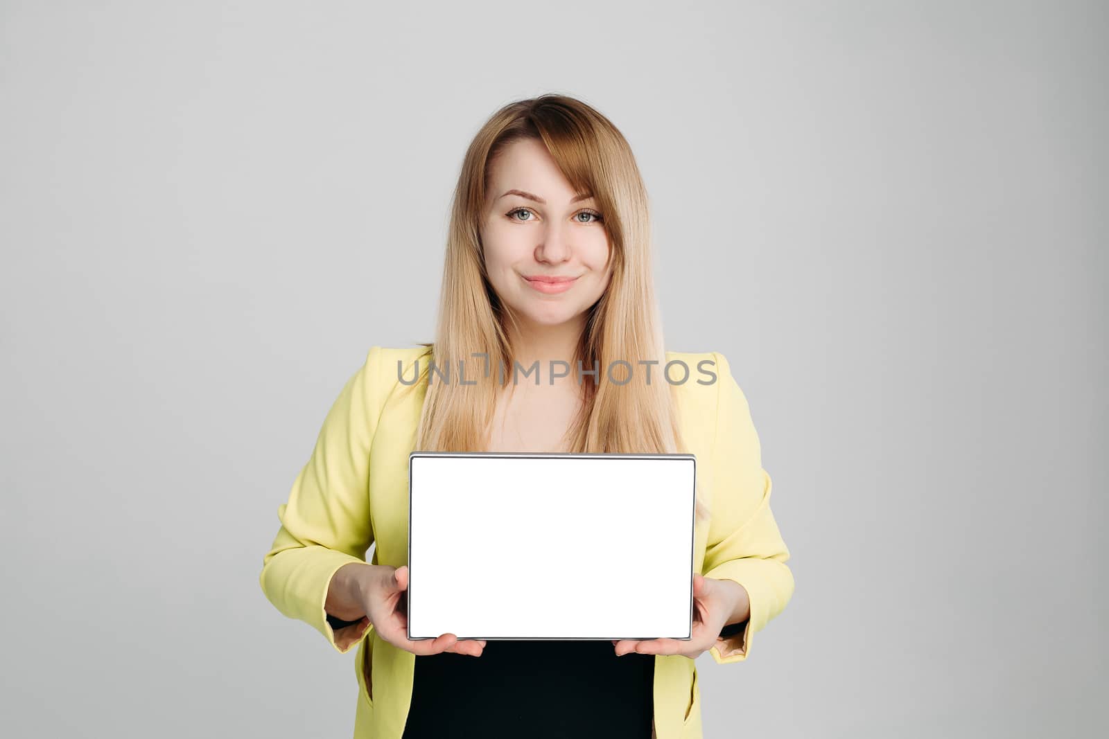 Portrait of blonde haired woman, wearing in yellow jacket and black blouse, holding tablet computer and showing at camera. Positivity girl posing against gray studio background. Concept of business.