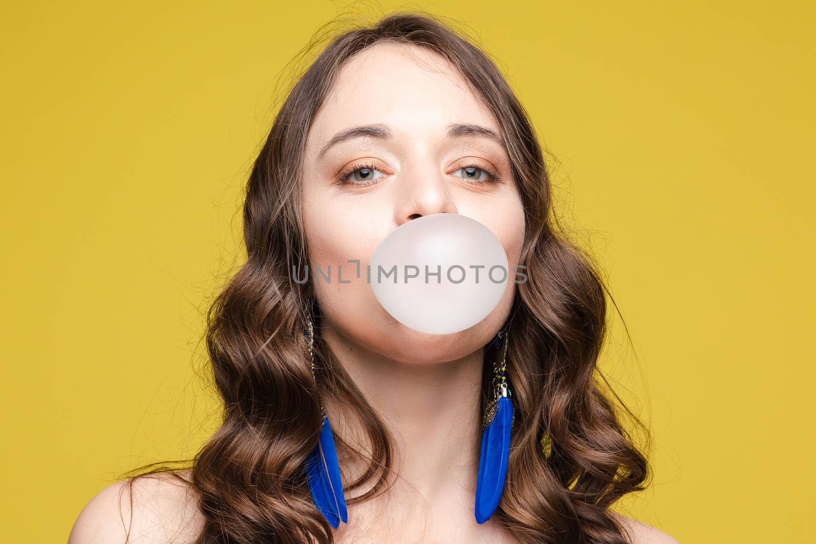 Front view of stylish female teenager wearing tracksuit and hat chewing gum and keeping big blue balloon in studio. Young girl looking at camera and posing on grey isolated background.