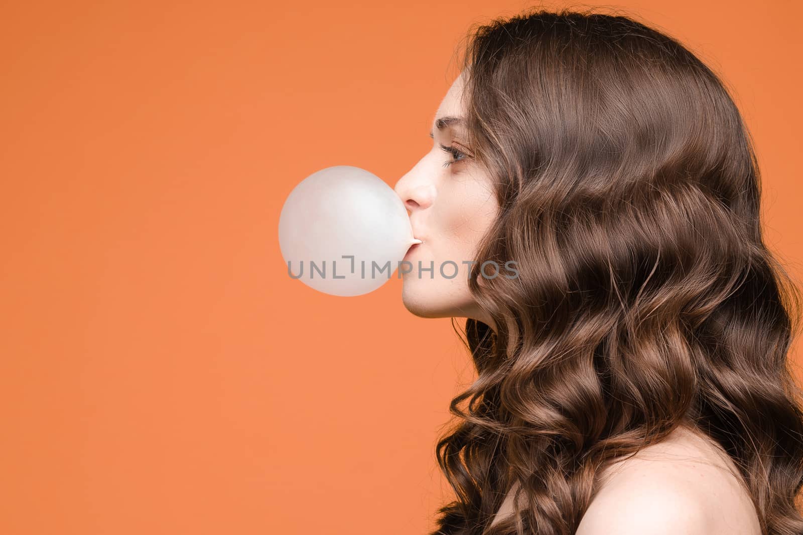 Front view of stylish female teenager wearing tracksuit and hat chewing gum and keeping big blue balloon in studio. Young girl looking at camera and posing on grey isolated background.