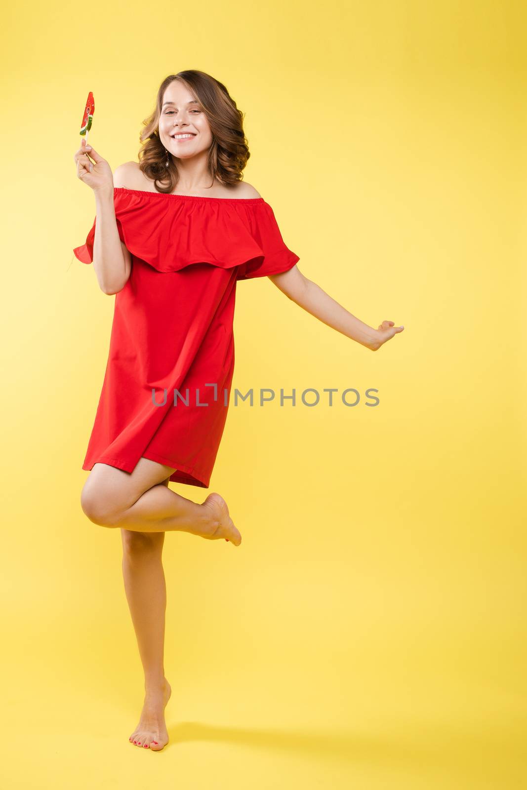 Beautiful and seductive woman wearing like sweet doll, posing at studio with candy on stick. Confident pretty woman in dress andsunglasses, holding hand on waist. Fashion, glamour.