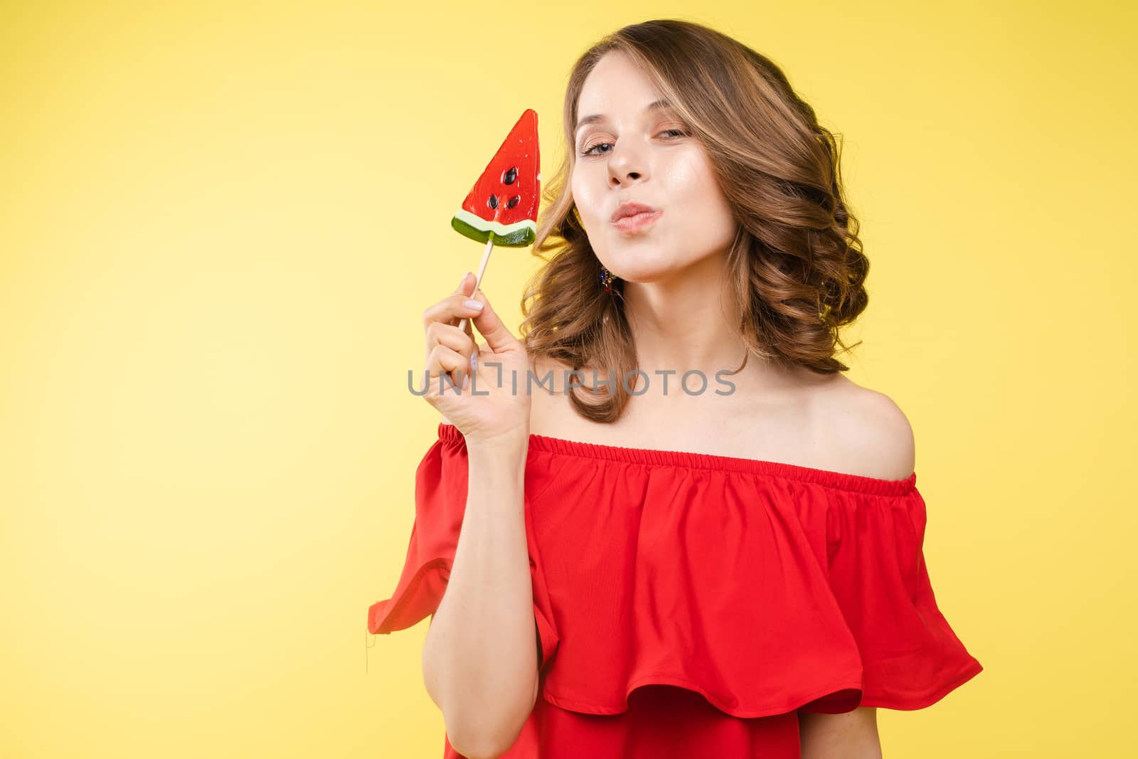 Close up portrait of attractive lovely girl in light dress handing lolipop isolated on yellow background