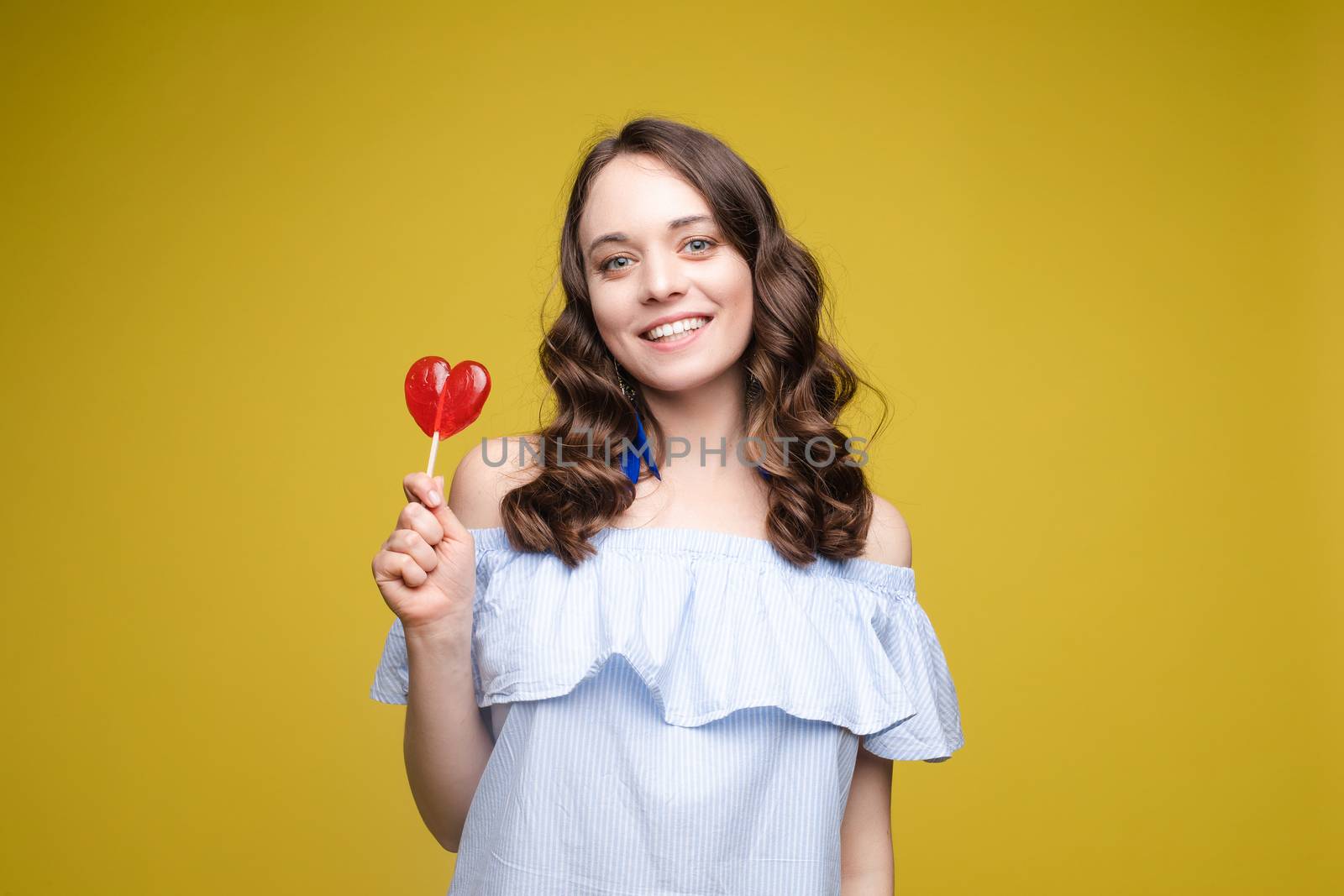 Beautiful and seductive woman wearing like sweet doll, posing at studio with candy on stick. Confident pretty woman in dress andsunglasses, holding hand on waist. Fashion, glamour.