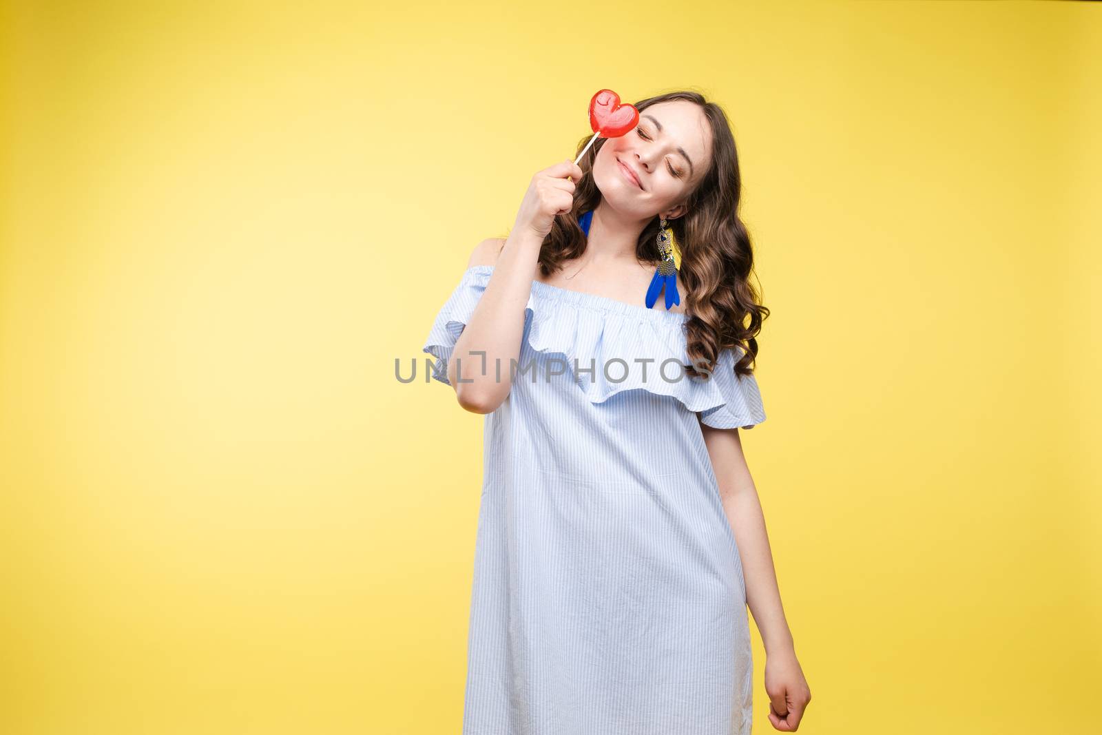 Beautiful and seductive woman wearing like sweet doll, posing at studio with candy on stick. Confident pretty woman in dress andsunglasses, holding hand on waist. Fashion, glamour.