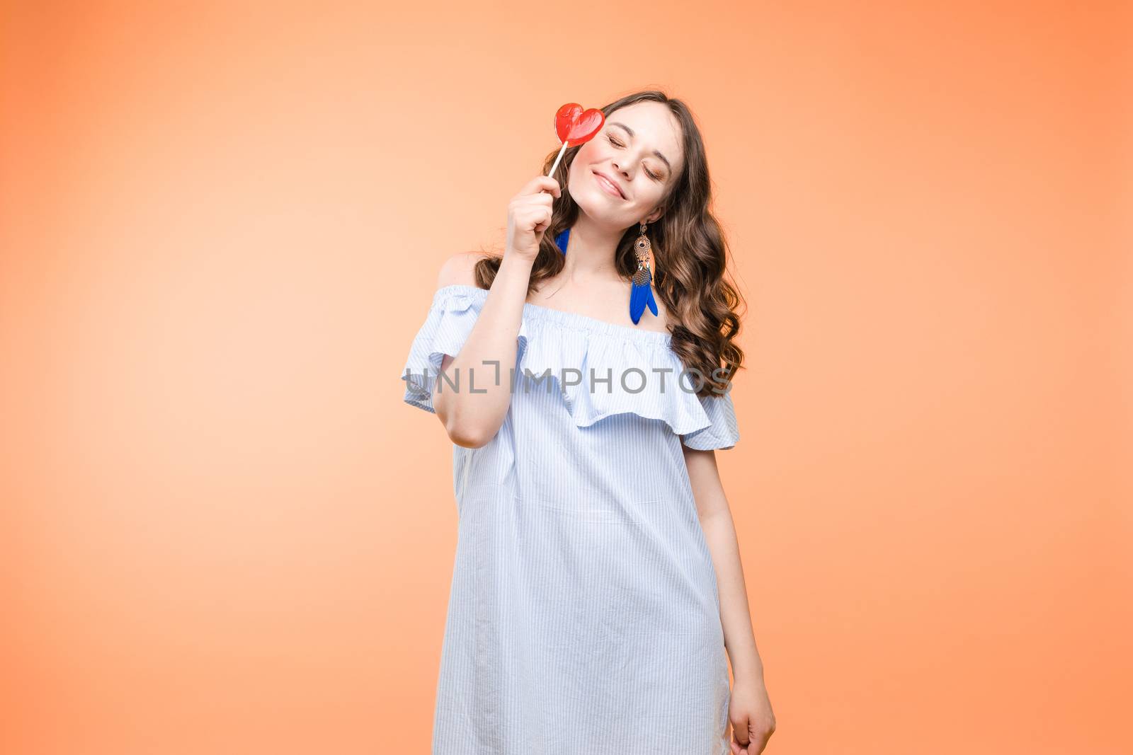 Beautiful and seductive woman wearing like sweet doll, posing at studio with candy on stick. Confident pretty woman in dress andsunglasses, holding hand on waist. Fashion, glamour.