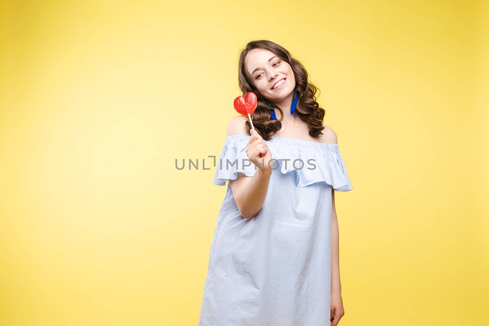 Beautiful and seductive woman wearing like sweet doll, posing at studio with candy on stick. Confident pretty woman in dress andsunglasses, holding hand on waist. Fashion, glamour.