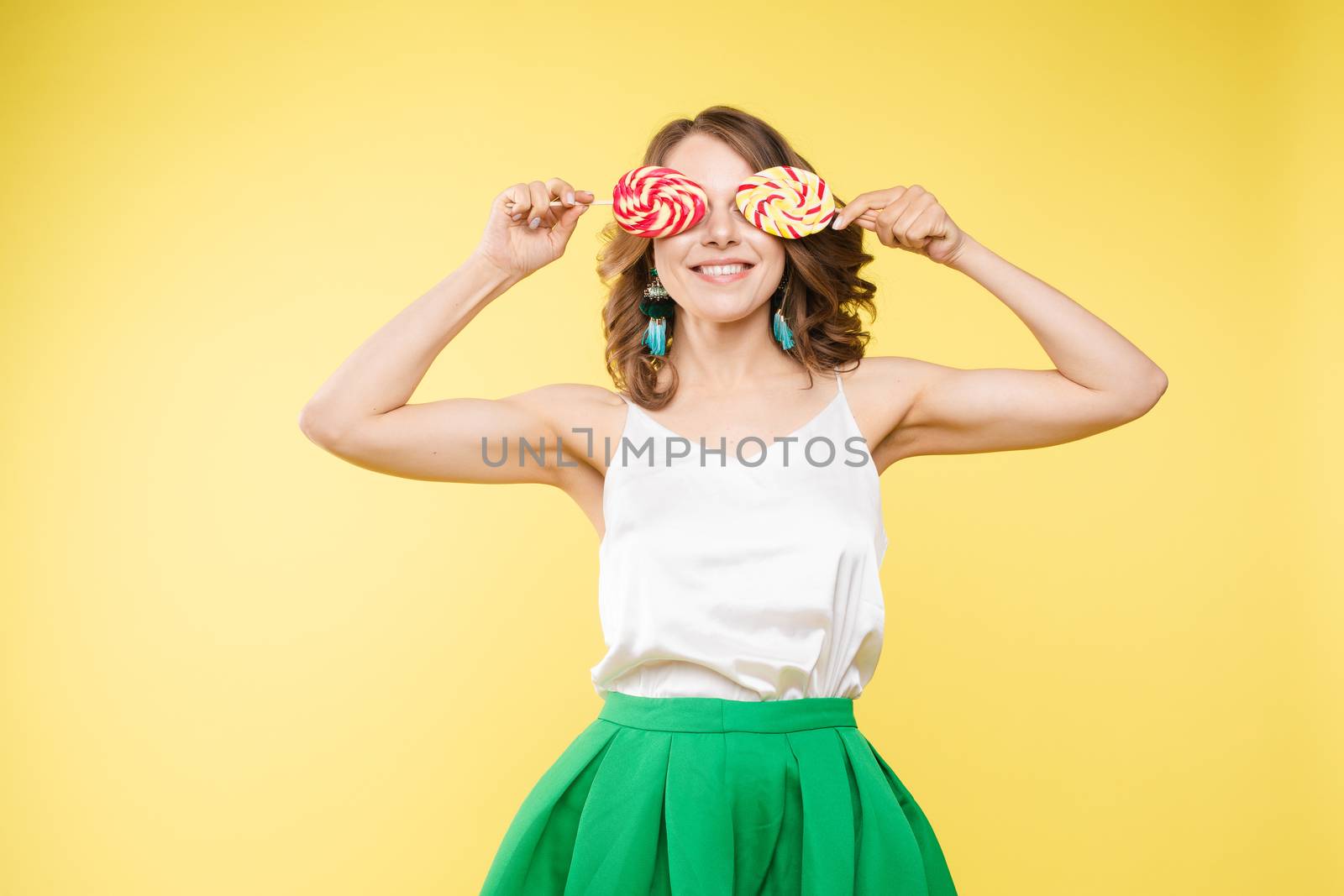 Full length studio portrait of laughing model in white top and green skirt and heels holding two sweet candies on her eyes like sunglasses with her mouth open. Isolate on yellow.