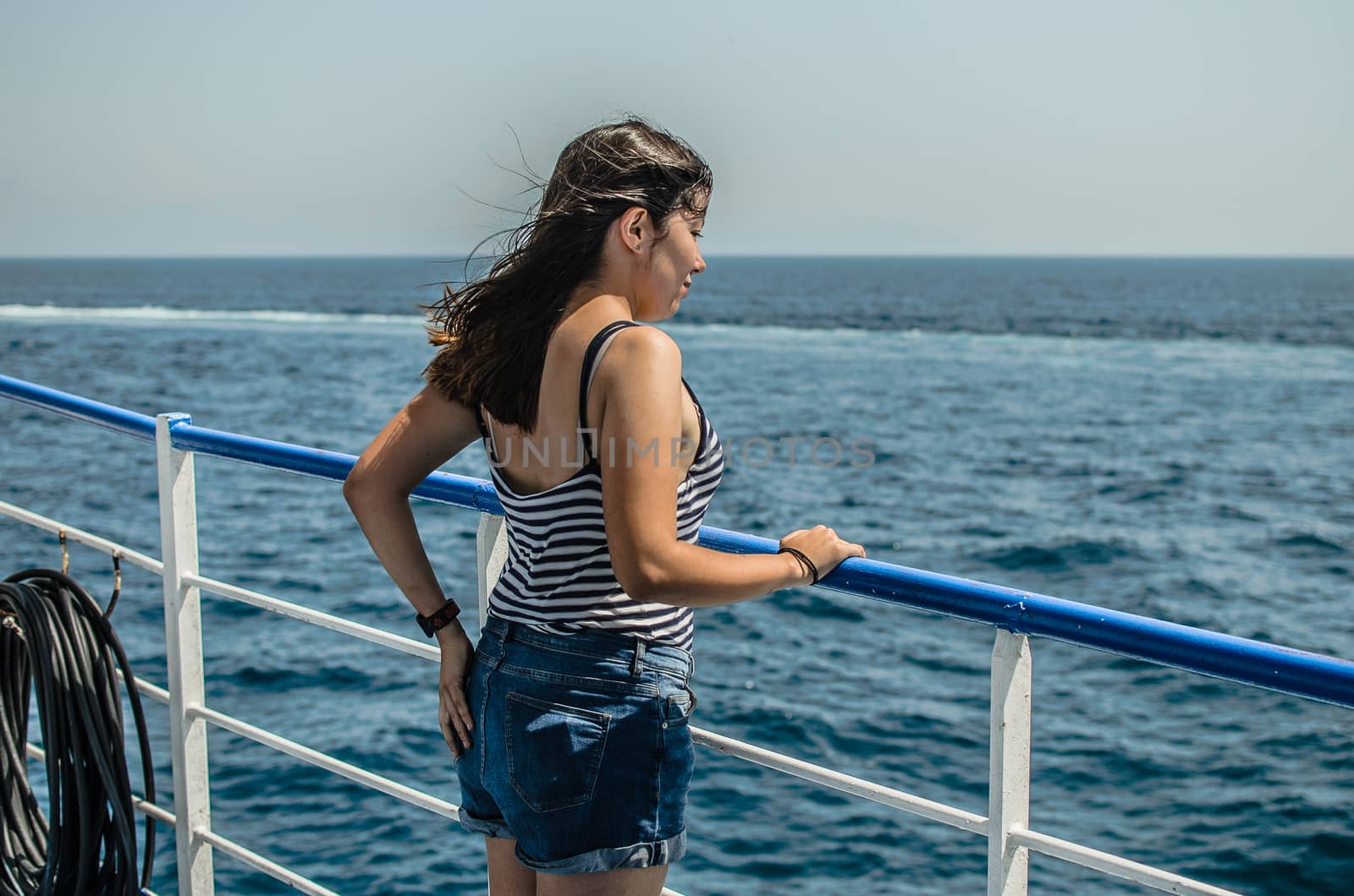 beautiful young girl with black hair in a striped t-shirt and blue denim shorts stands on the deck of a ship at sea