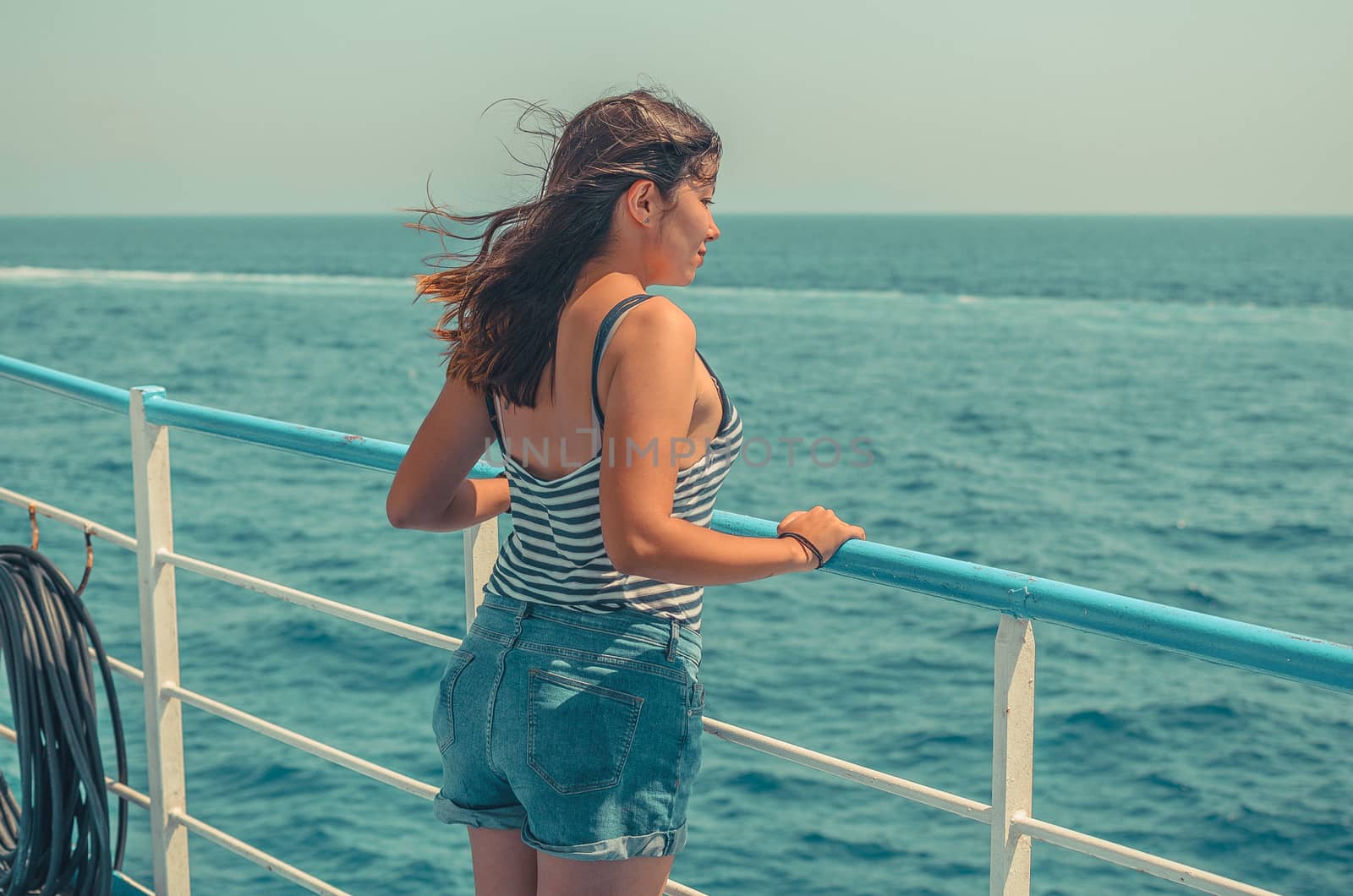 beautiful young girl with dark hair in a striped t-shirt and blue denim shorts stands on the deck of a ship at sea