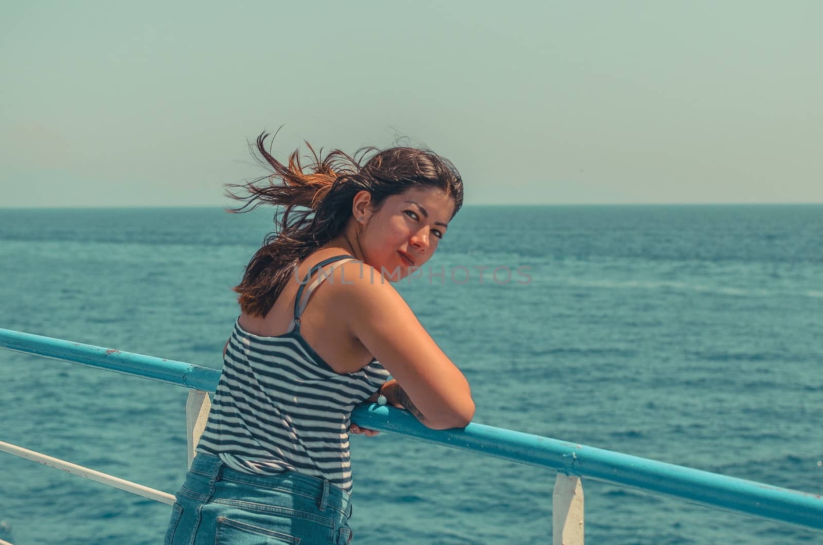 cheerful young girl with black hair in a striped t-shirt and blue jeans stands on the deck of a ship at sea by chernobrovin