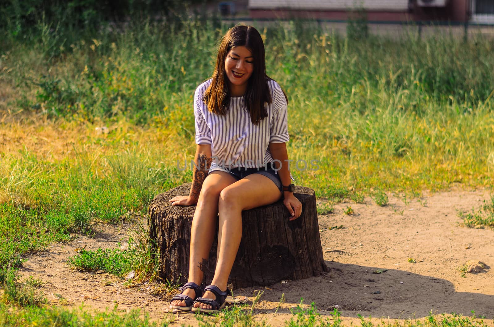 A beautiful, brunette woman in a white shirt and blue denim shorts sits on a stump in the park by chernobrovin