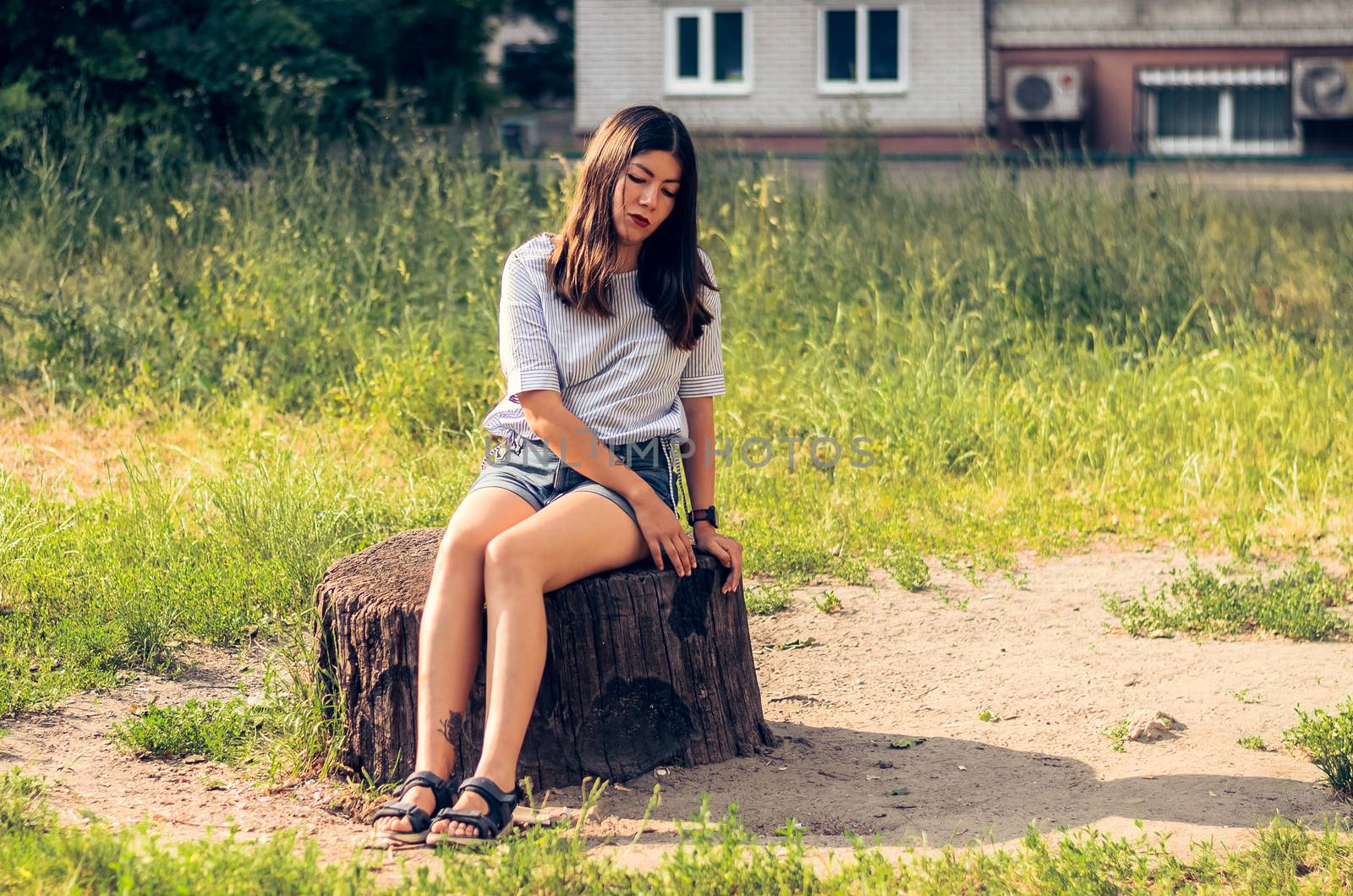 a girl with black hair, dressed in a white blouse and blue denim shorts, sits on a stump