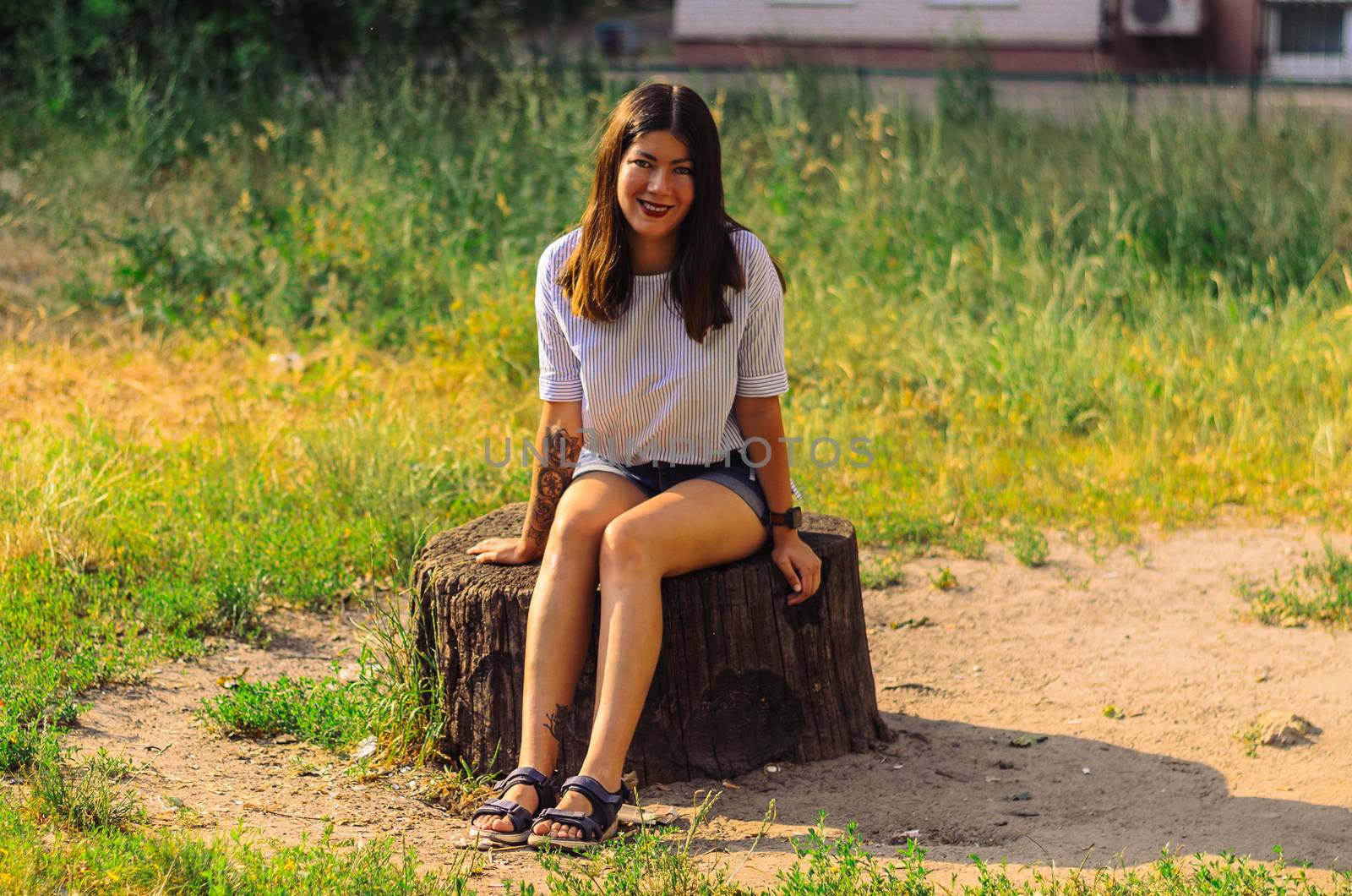 A beautiful, brunette girl in a white shirt and blue jeans shorts sits on a stump in the park