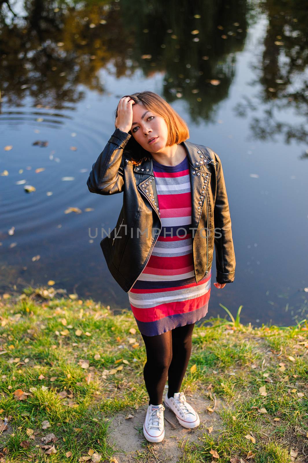 lovely red-haired young girl in a black leather jacket, striped sweater and white gym shoes on a green shore