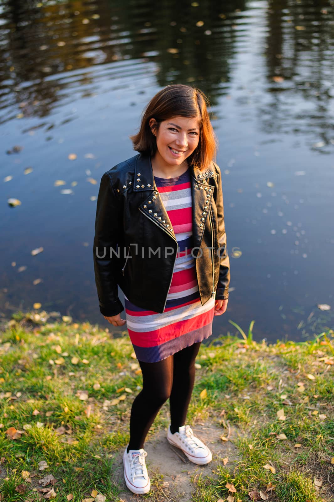 pretty fashionable young woman in a colorful striped sweater and black leather jacket stands on the shore