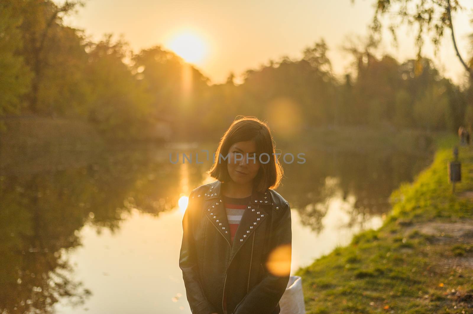 young brunette girl in a black jacket and striped multi-colored t-shirt stands on a background of a forest lake at sunset