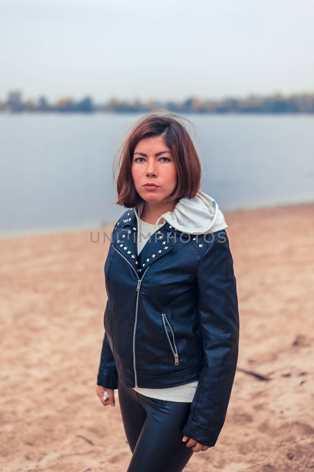 young pretty girl in a black jacket stands on a sandy beach near the water