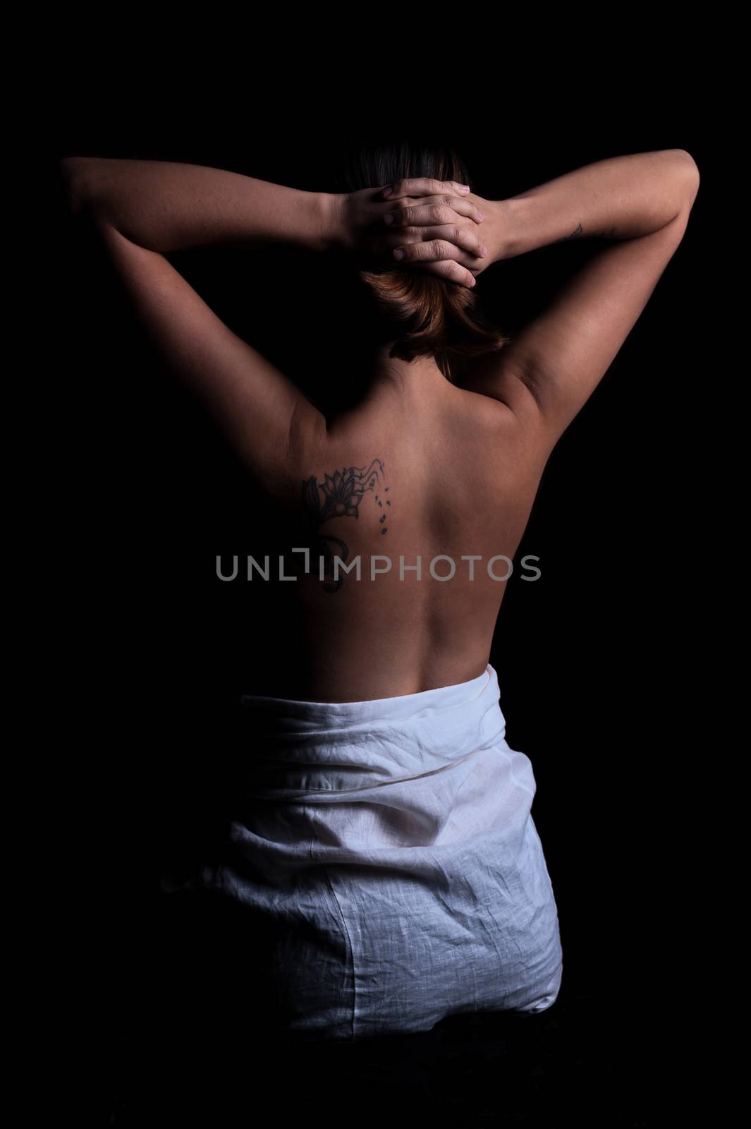 young girl with dark hair and a tattoo stands with naked back in the dark, holding her hands on her head