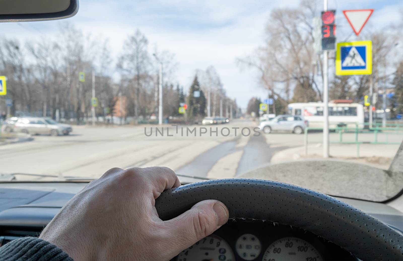 the driver hand on the steering wheel of a car located in front of a red prohibiting traffic light, pedestrian crosswalk, and cars that the driver gives way to
